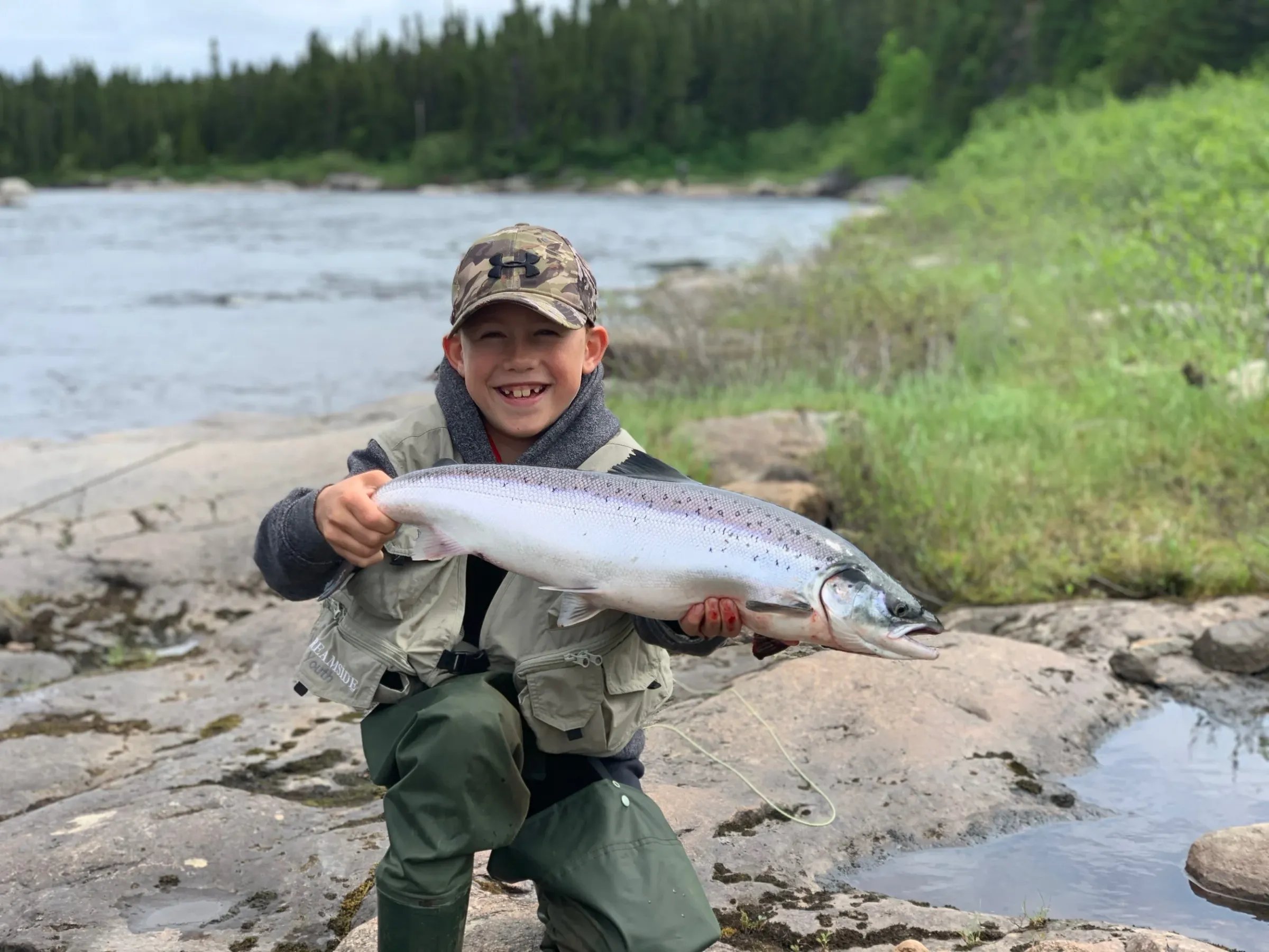 Young boy smiling and holding a large Atlantic salmon on a rocky riverbank