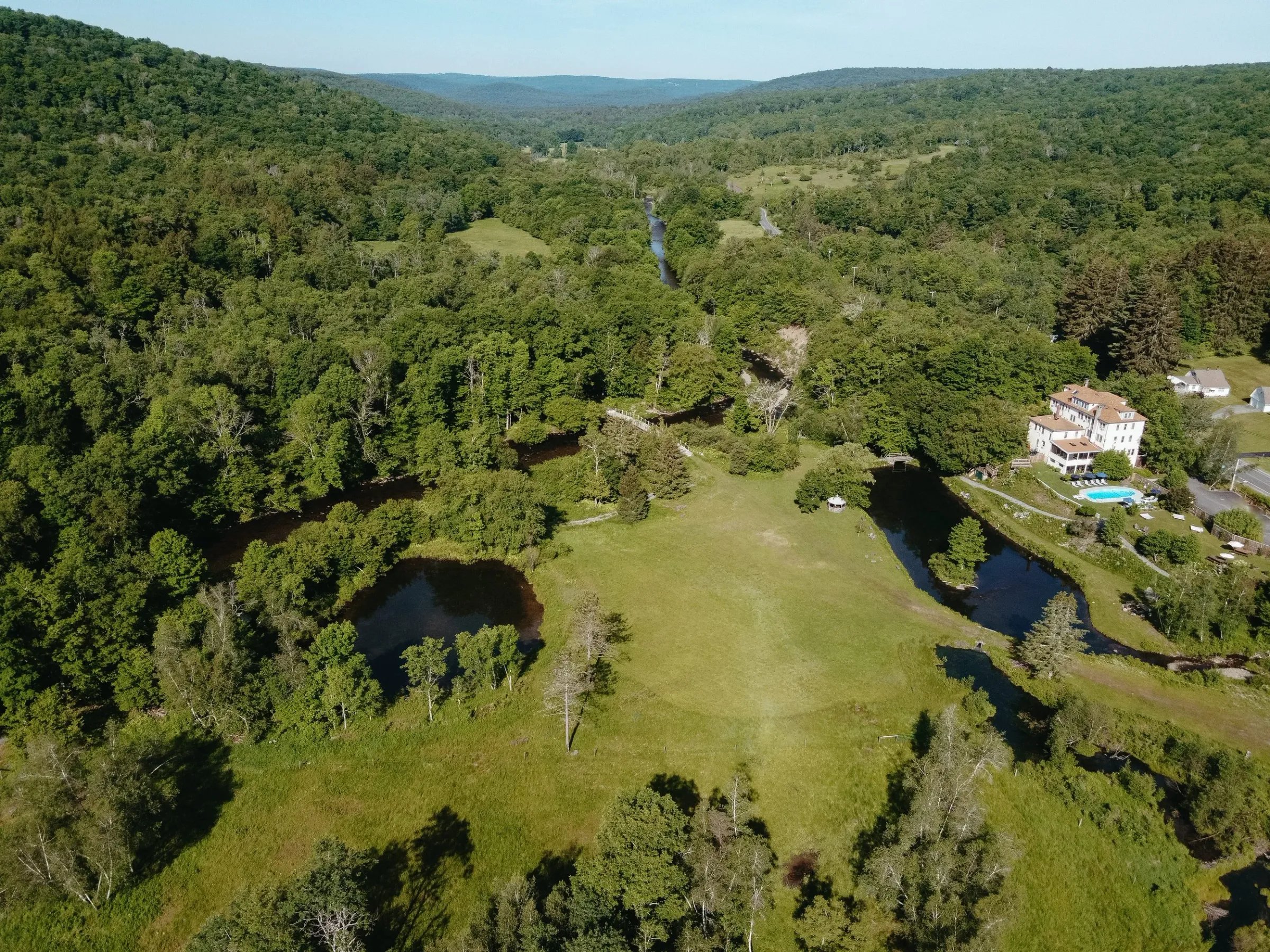 Aerial view of a countryside estate with large house, swimming pool, pond, and lush green trees surrounded by rolling hills.