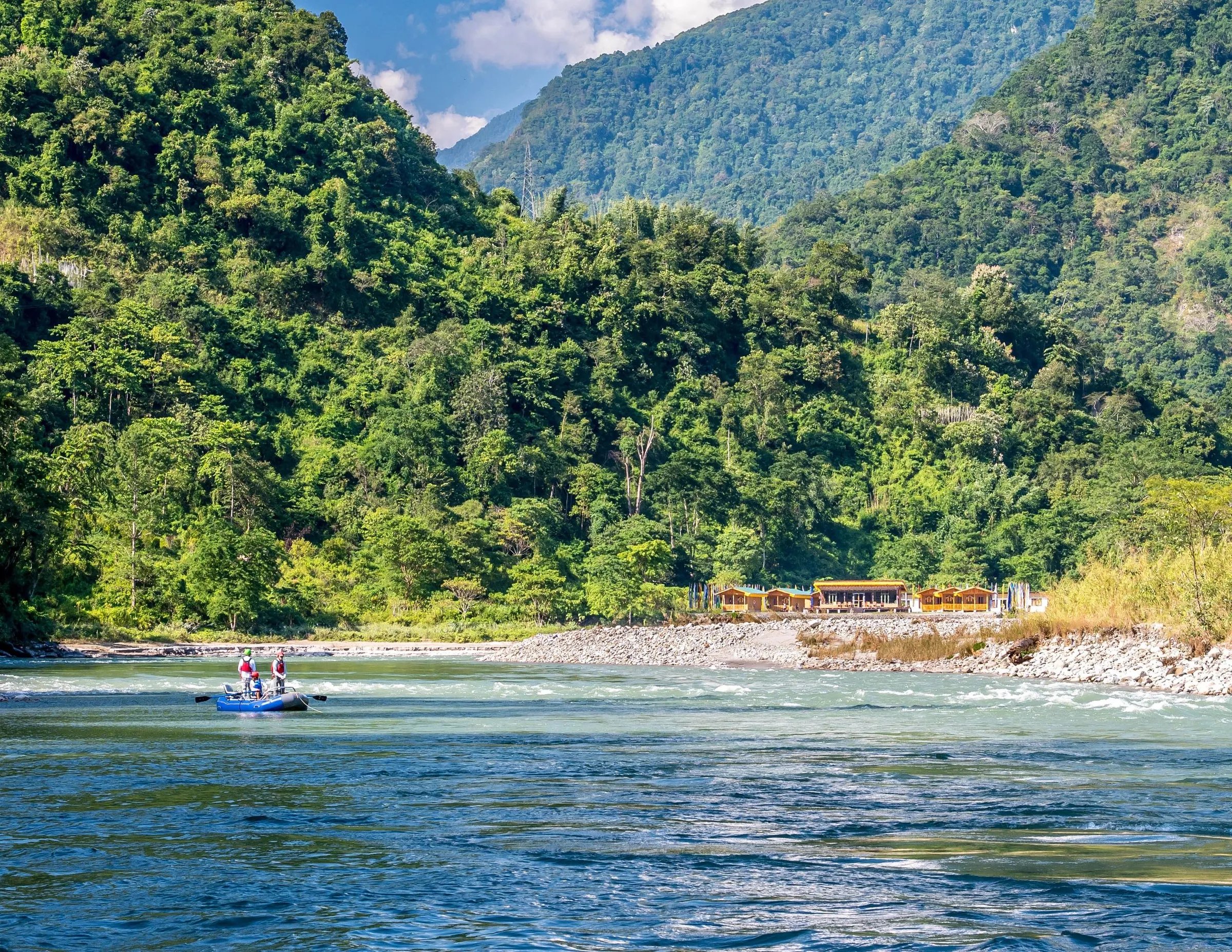 Raft on a wide river with lodge buildings nestled on the far bank against mountains