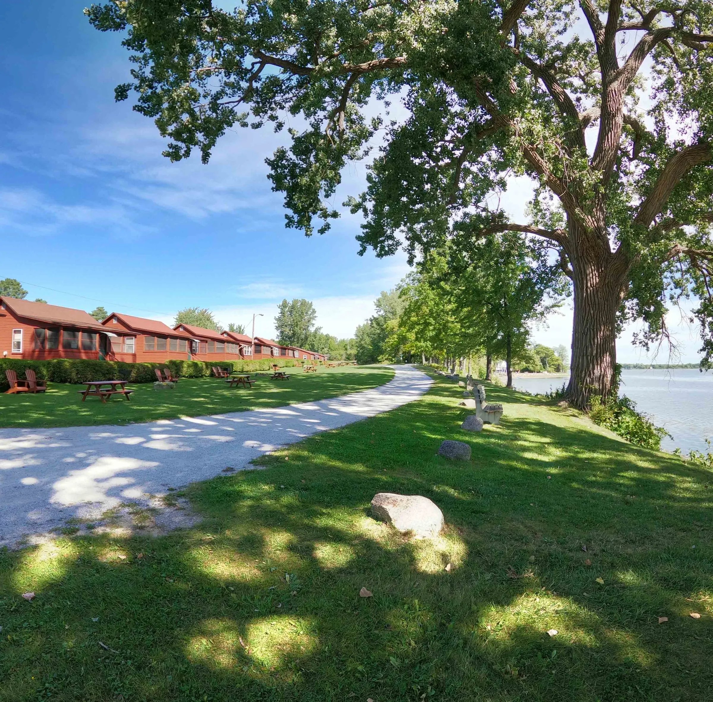 Grassy grounds with a gravel path, large trees, and a row of red cottages beside a lake