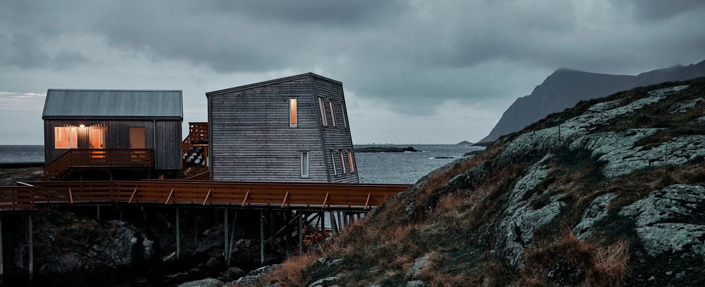 Modern and traditional lodge buildings on a rocky coastline under a moody sky.