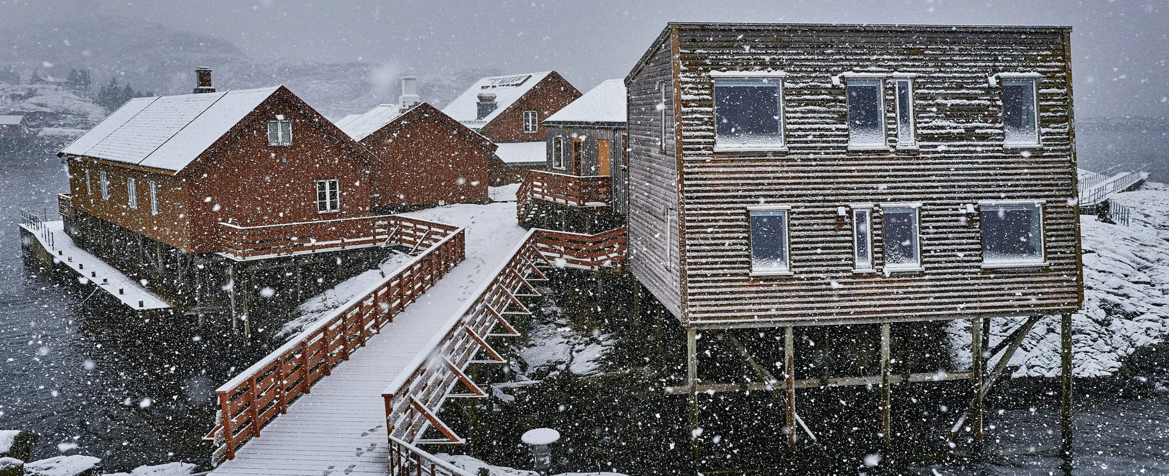 Snow-covered lodge buildings on stilts over the water during a snowfall.