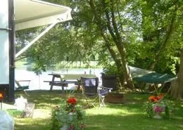 RV parked at a lakeside campsite with a picnic table and chairs under trees.