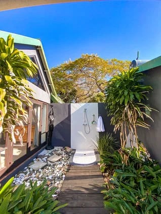 Outdoor shower area with white wall and wooden path, surrounded by tropical plants
