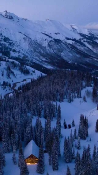 Aerial view of a rustic lodge nestled in a snowy forest at dusk, surrounded by mountains