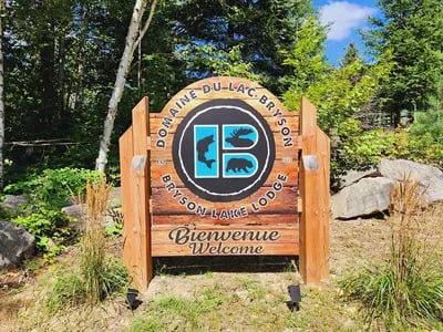 Wooden welcome sign for Domaine du Lac Bryson / Bryson Lake Lodge in a wooded setting