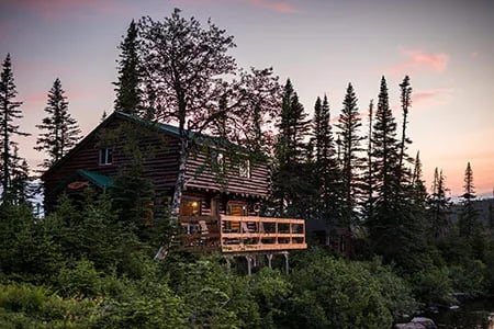 Wooden lodge with a green roof and a deck, surrounded by trees at dusk.