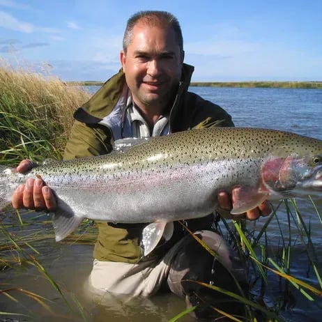 Man holding a large rainbow trout in a river with tall grass.