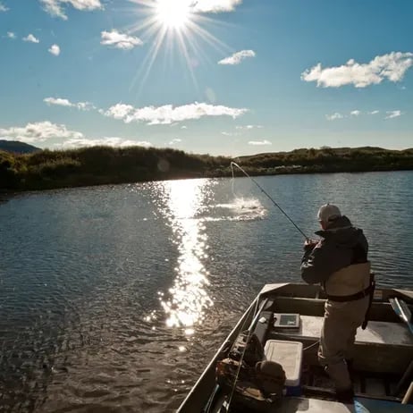 Angler casting from a drift boat with a fish jumping in the sunlit water.