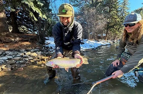 Two anglers in a snowy river, one holding a large rainbow trout