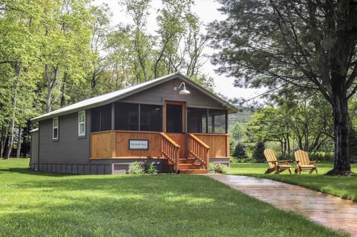 Small gray cabin with wooden porch and steps on green lawn surrounded by trees, with two wooden chairs near a large tree.