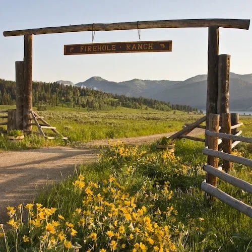 Wooden entrance gate with "Firehole Ranch" sign, yellow wildflowers, and mountains in the background.