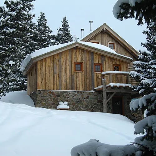 Wooden hut with a stone foundation covered in snow, surrounded by pine trees.