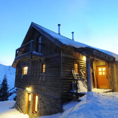 Wooden hut with glowing windows at dusk, surrounded by deep snow.