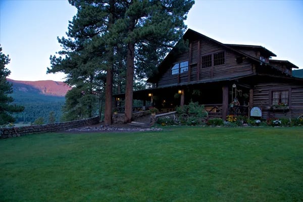 Main lodge building at dusk, nestled among tall pine trees with mountains in the distance