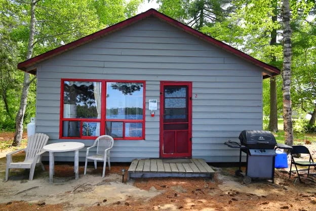 Front exterior of a grey cabin with red trim, a small deck, and a grill