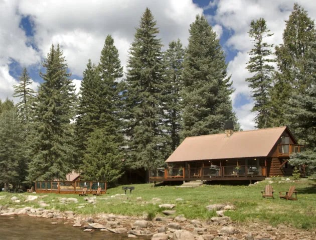 Log cabin by a river surrounded by tall pine trees on a sunny day