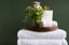 A wooden tray holding a rolled-up white towel and a glass vase with assorted fresh flowers, placed on top of two folded white towels, with a guest information card behind the arrangement, against a dark green wall.