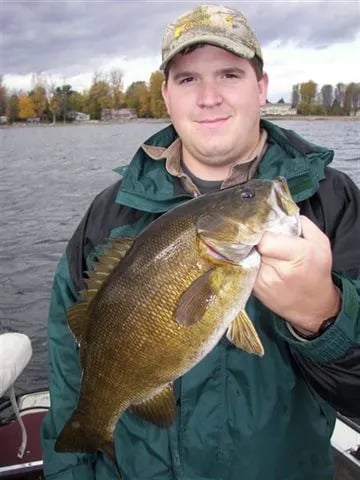 Man in sunglasses and cap holding a smallmouth bass against a blue sky over water.