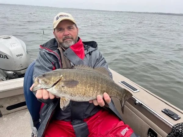 Smiling man in a hat holding a large smallmouth bass with a wooded shoreline behind him.