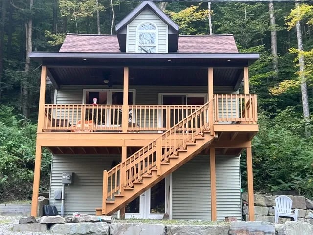 Two-story lodge with a large wooden deck and stairs, nestled in a green forest.