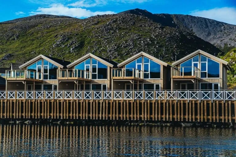 Row of modern waterfront cabins with large windows and balconies, reflecting in calm water under a blue sky