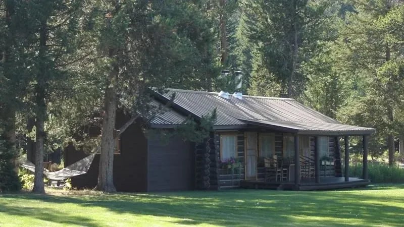 Rustic cabin exterior with a covered porch, surrounded by green grass and tall trees.
