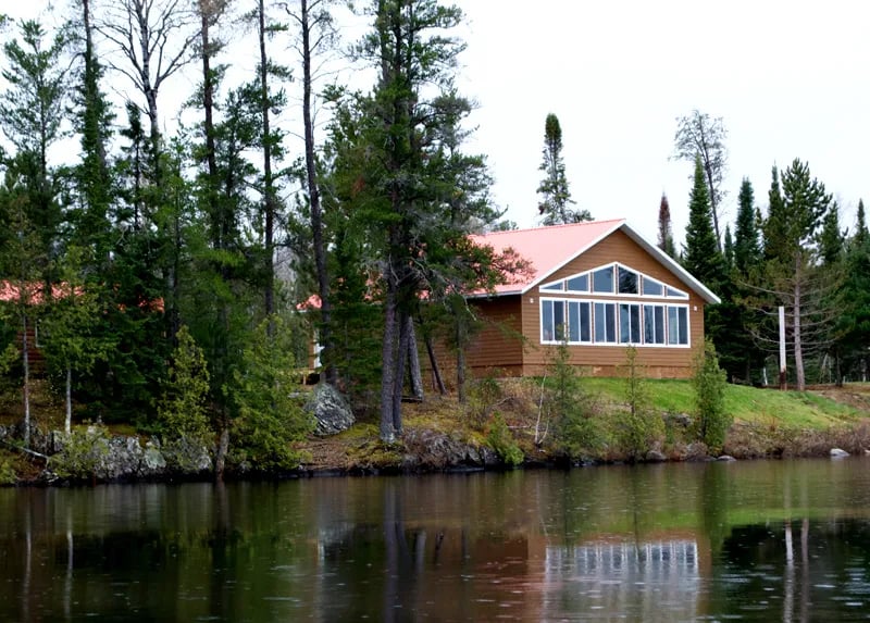 Rustic lodge building with a red roof and large windows, situated on a lakefront