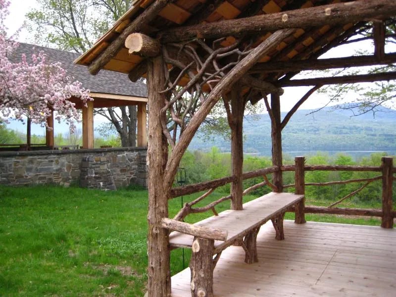 Rustic wooden gazebo surrounded by flowering crabapple trees with a lake in the background under a blue sky.