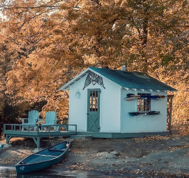 Charming lakeside boathouse with Adirondack chairs, canoe, and kayaks, set against vibrant autumn foliage.