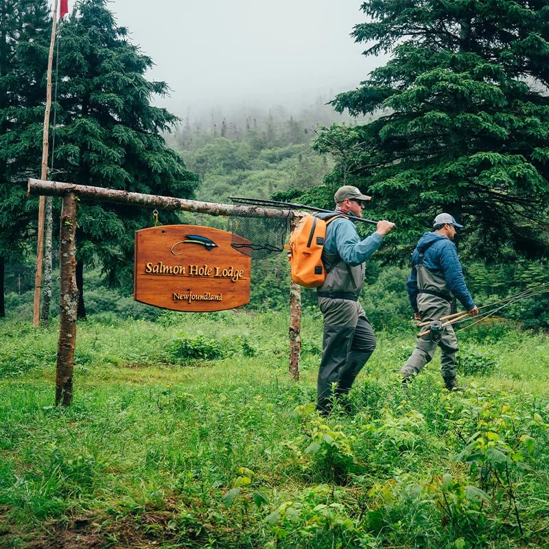 Two anglers walking past a rustic wooden Salmon Hole Lodge sign in a lush forest