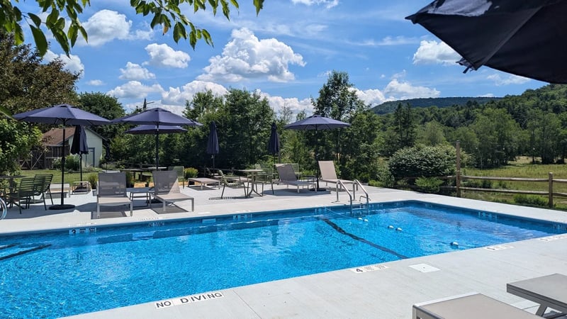 Empty outdoor swimming pool with several lounge chairs and umbrellas, surrounded by trees and mountains under a partly cloudy sky.