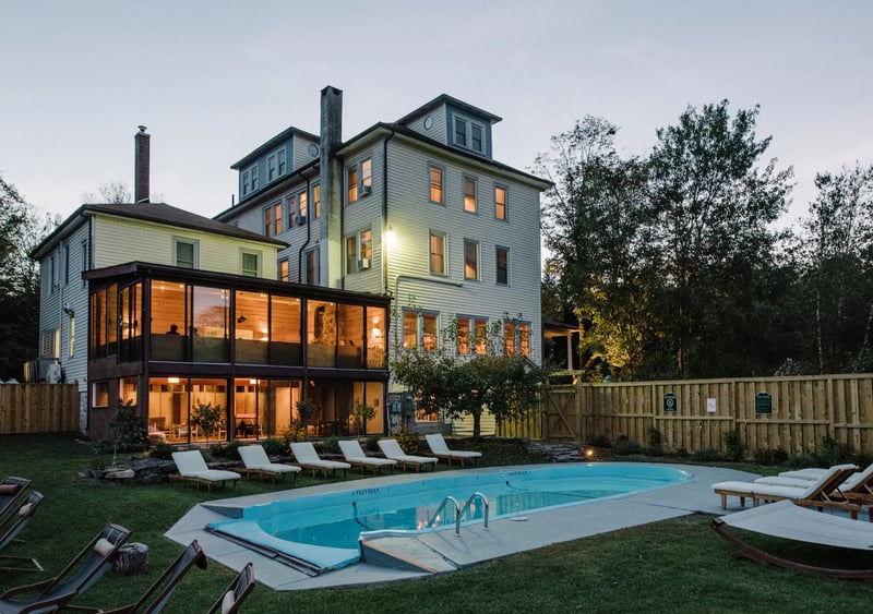A large multi-story house with an outdoor pool and a fenced backyard at dusk. The house has lit windows and a glass-enclosed porch, with lounge chairs around the pool.