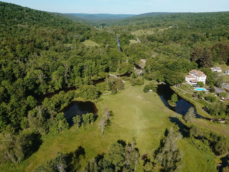 Aerial view of a countryside estate with large house, swimming pool, pond, and lush green trees surrounded by rolling hills.