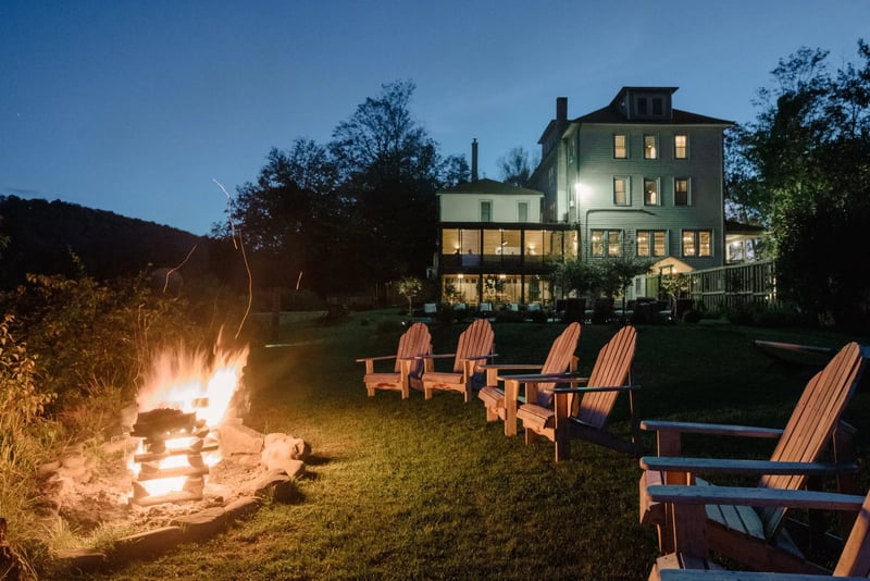 Nighttime backyard scene with a lit campfire on the left side, surrounded by wooden Adirondack chairs, with a large multi-story house in the background. The house's interior lights are visible through the windows, and trees are on either side. The sky is dark blue.