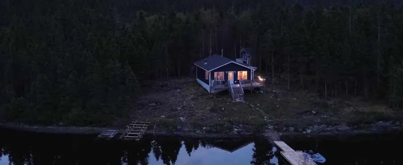 Cozy cabin with lit windows by a dark forest and calm water at dusk.
