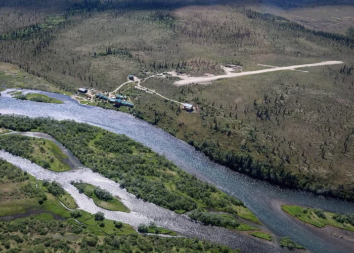 Main lodge building and several small cabins with green roofs along a riverbank under a cloudy sky