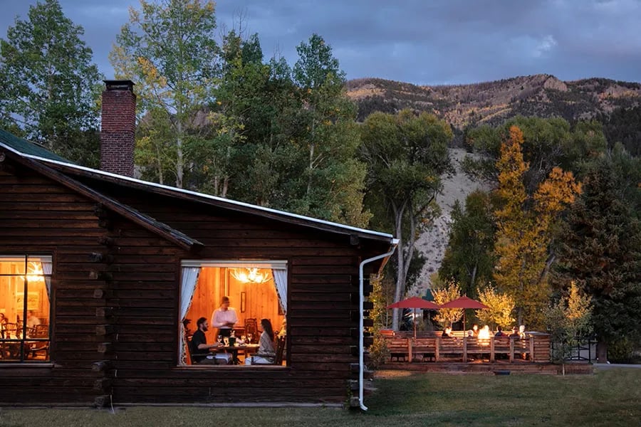 Side view of a log lodge with illuminated windows and an outdoor dining deck at dusk.