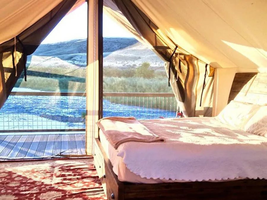 Glamping tent interior with a comfortable bed and large window overlooking a river and hills.