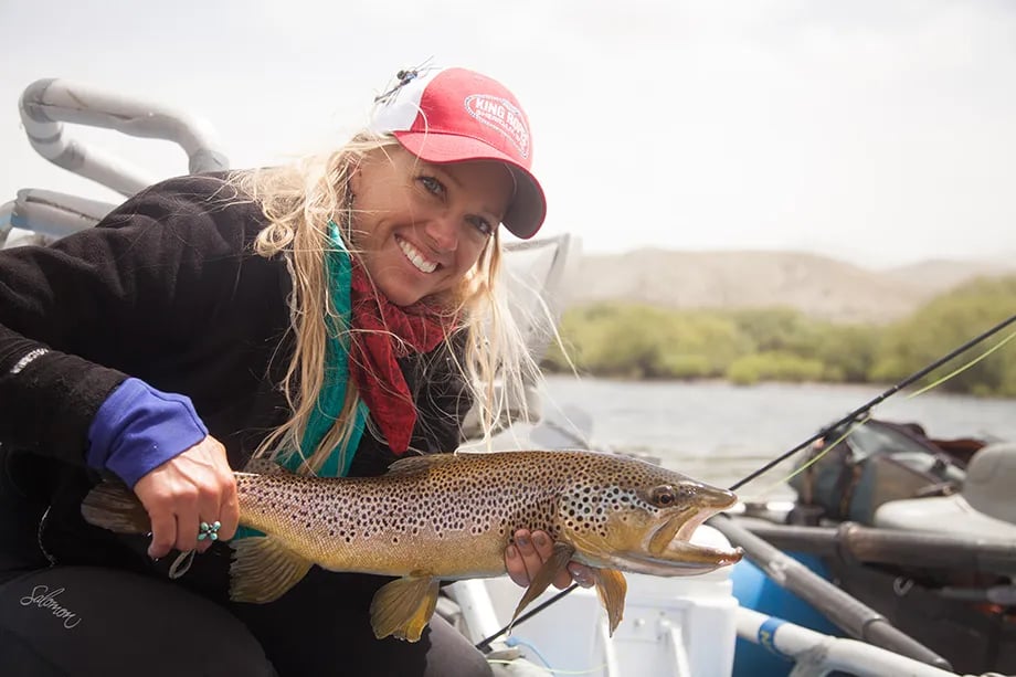 Woman smiling, holding a large spotted brown trout on a river.