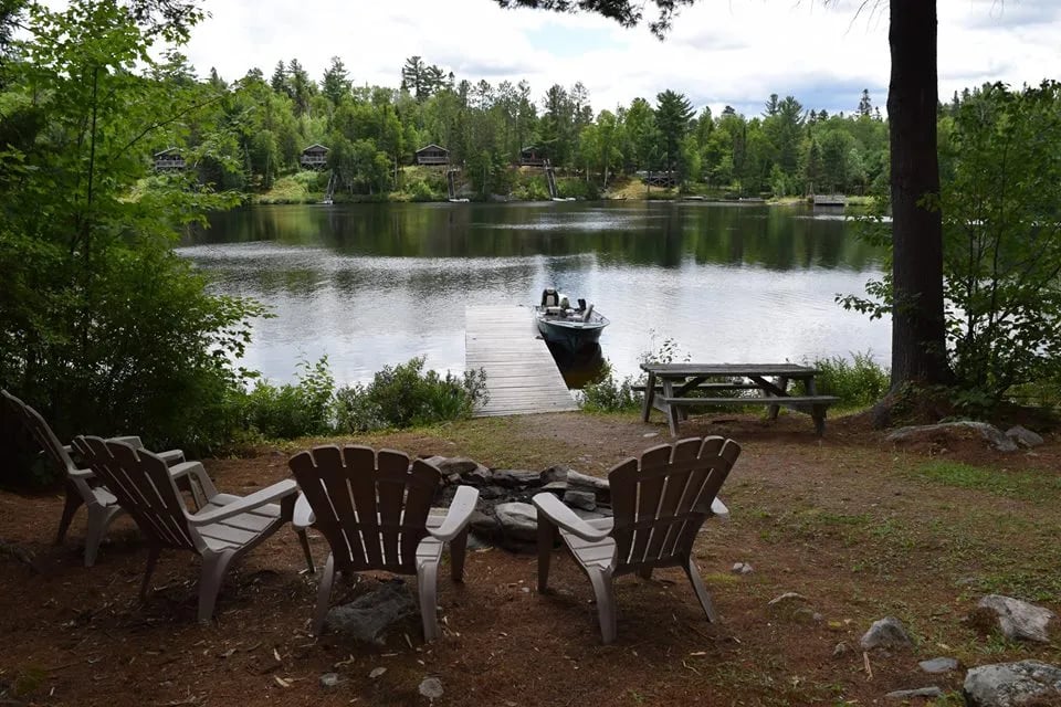 Lakeside fire pit area with Adirondack chairs and a dock leading to a boat.
