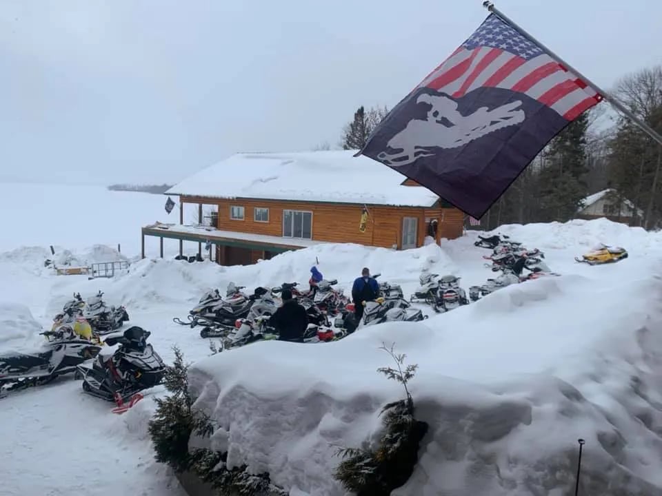 AJ's Lodge & Oven in winter, covered in snow, with snowmobiles parked outside and an American flag flying